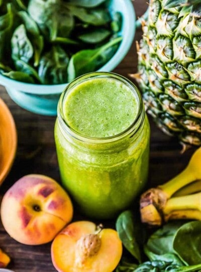 Green pre-workout smoothie in a glass jar surrounded by fresh spinach, bananas, peaches, and pineapple, perfect for a pre-workout smoothie recipe.