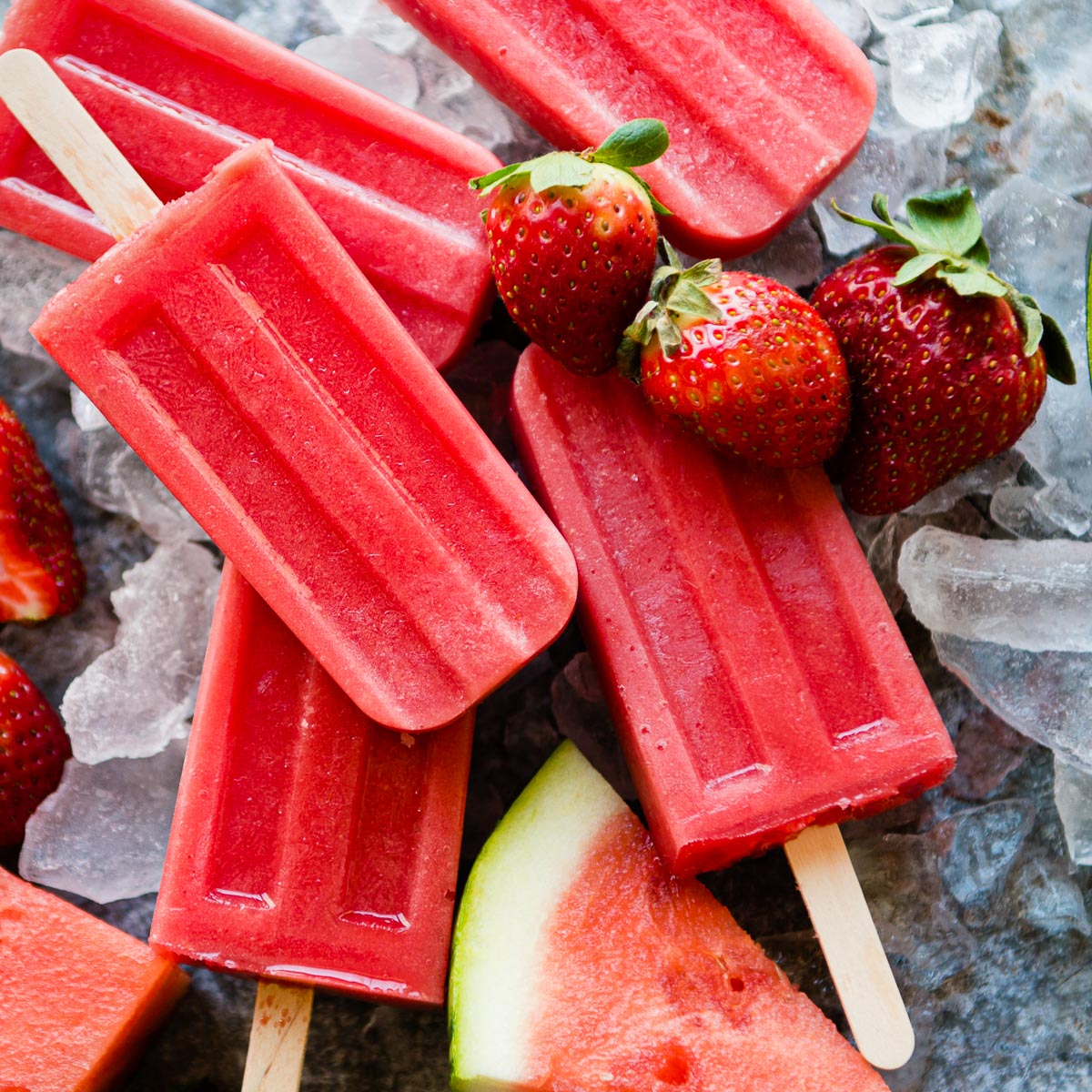 5 bright red popsicles surrounded by fruit and ice.