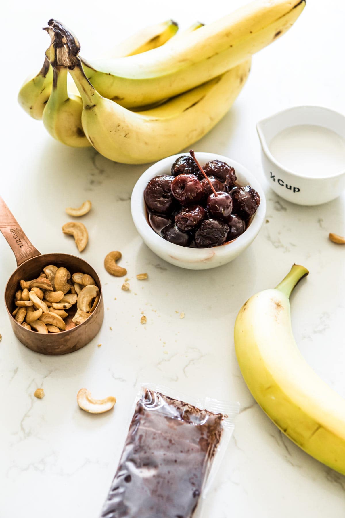 marble surface with bananas, frozen cherries, cashew milk, cashews, and a packet of açai puree.