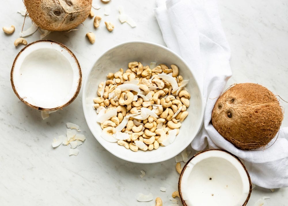 white bowl of raw cashews and coconut flakes, surrounded by fresh coconut halves and wholes.