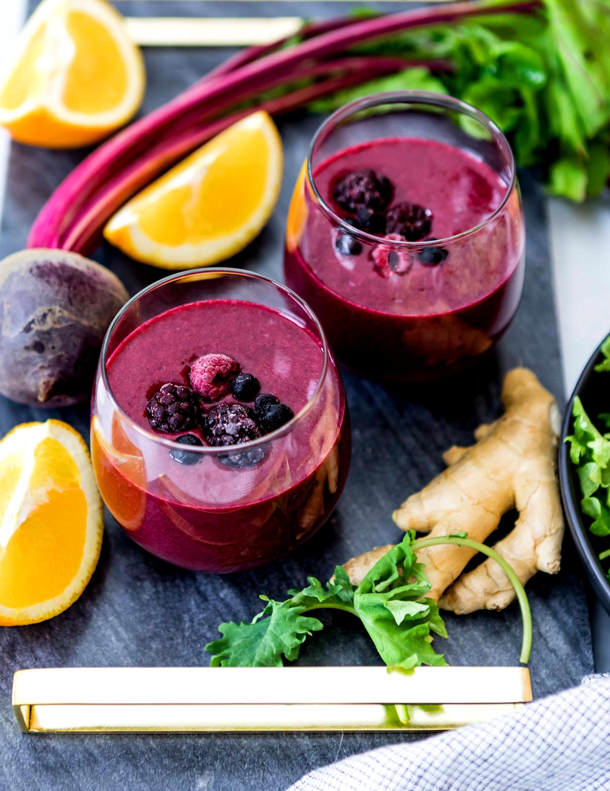 Two glasses full of one of the best smoothie recipes for anti-inflammatory, topped with berries on a countertop, surrounded by ginger root, kale lemon slices, and beets.