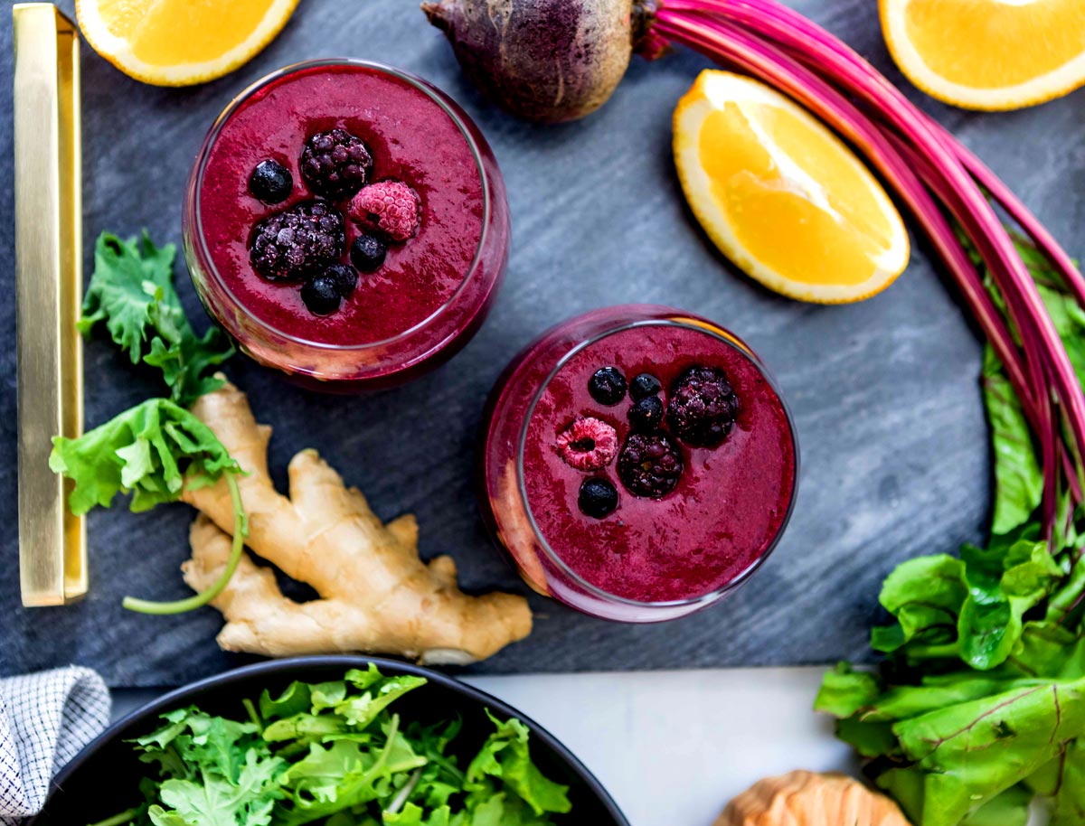 Two inflammatory berry smoothies, topped with berries on a countertop, surrounded by anti inflammatory ingredients such as lemon, beets, ginger and kale.