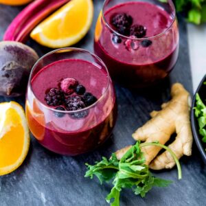 Two glasses full of one of the best smoothie recipes for anti-inflammatory, topped with berries on a countertop, surrounded by ginger root, kale lemon slices, and beets.