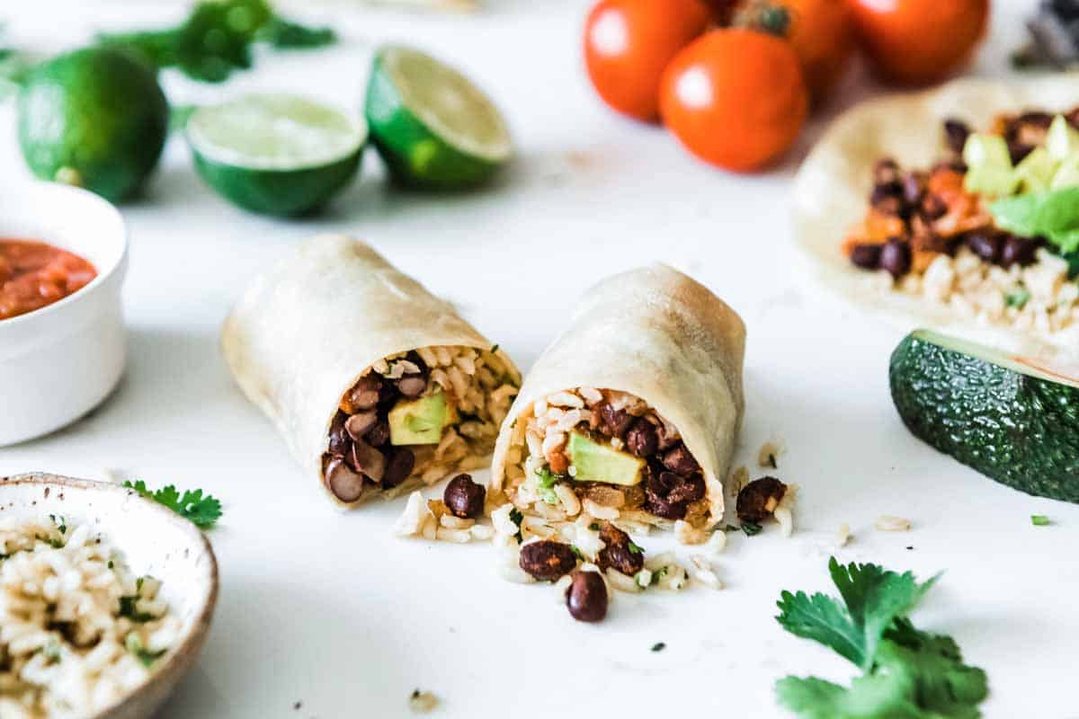 bean and rice burritos cut open to show the ingredients inside on a white counter surrounded by fresh ingredients.