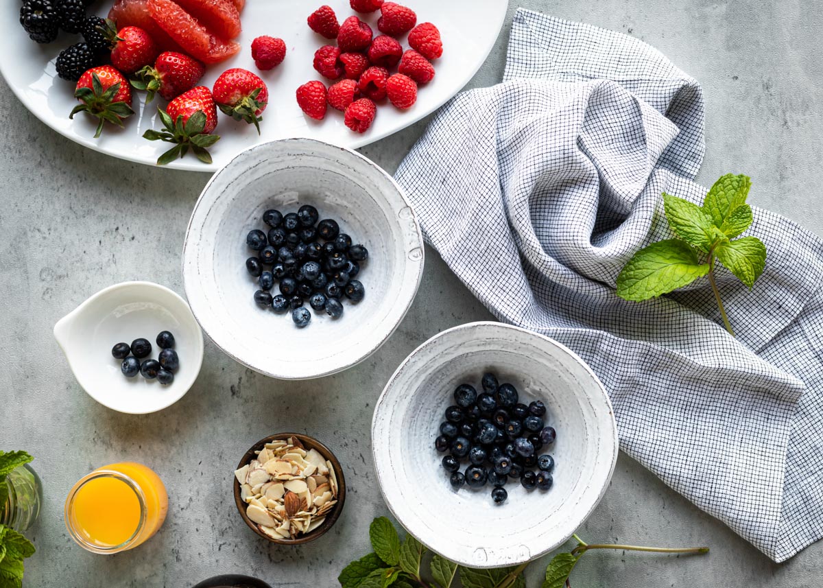 3 white bowls with blueberries in them as well as a bowl of almonds and some orange juice.