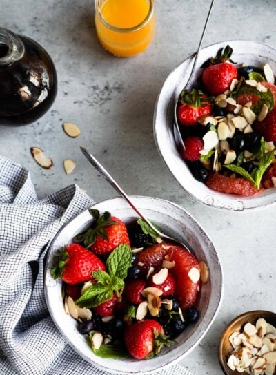 2 white bowls of a breakfast fruit salad on a table with orange juice, maple syrup and sliced almonds.