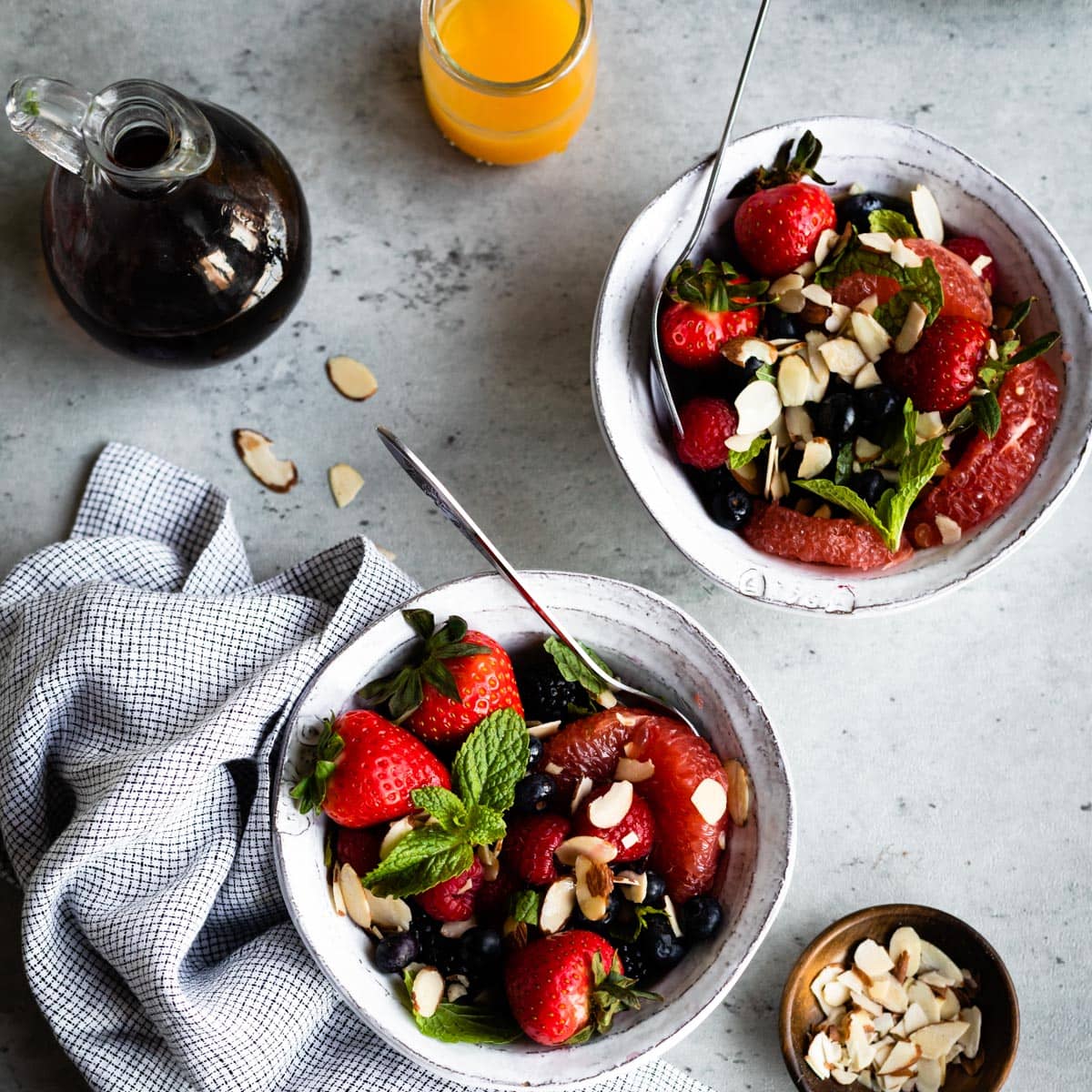 2 white bowls of a breakfast fruit salad on a table with orange juice, maple syrup and sliced almonds.