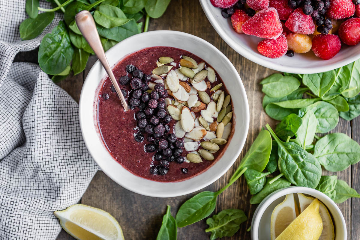 bright snack bowl topped with fresh fruit, nuts and seeds, surrounded by spinach, lemons and frozen fruit.