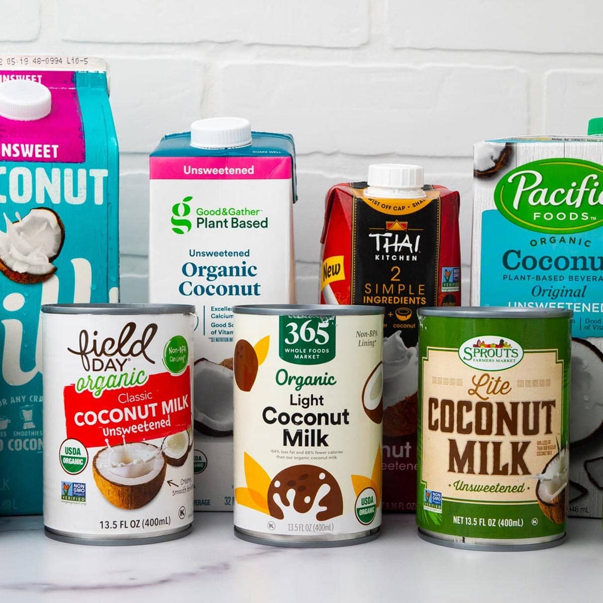 7 coconut milk brands grouped on a countertop with a white brick background.