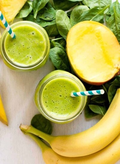 Healthy green smoothie in a glass jar with straw, placed on a countertop, with fruits and ingredients are blurred in the background.