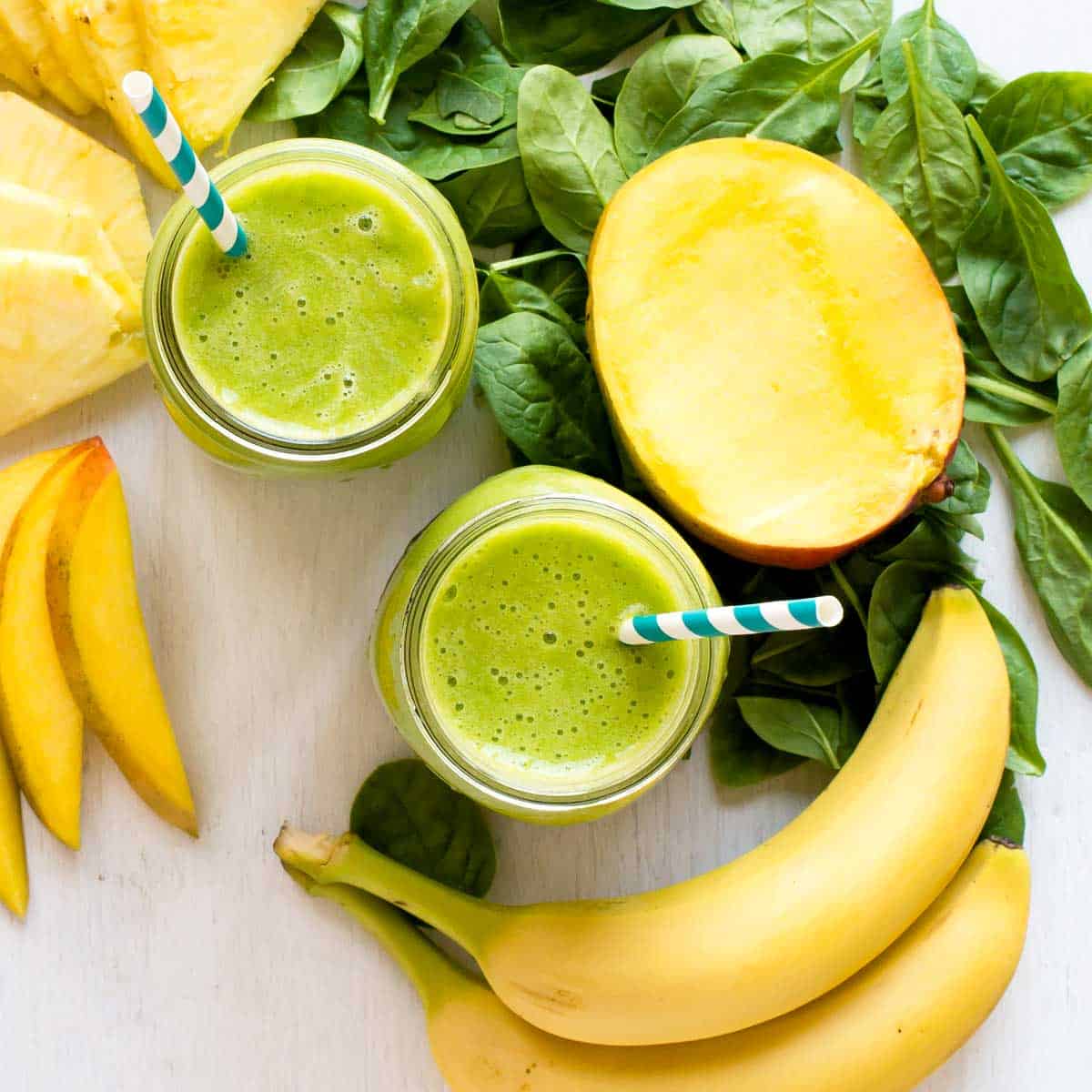 Healthy green smoothie in a glass jar with straw, placed on a countertop, with fruits and ingredients are blurred in the background.