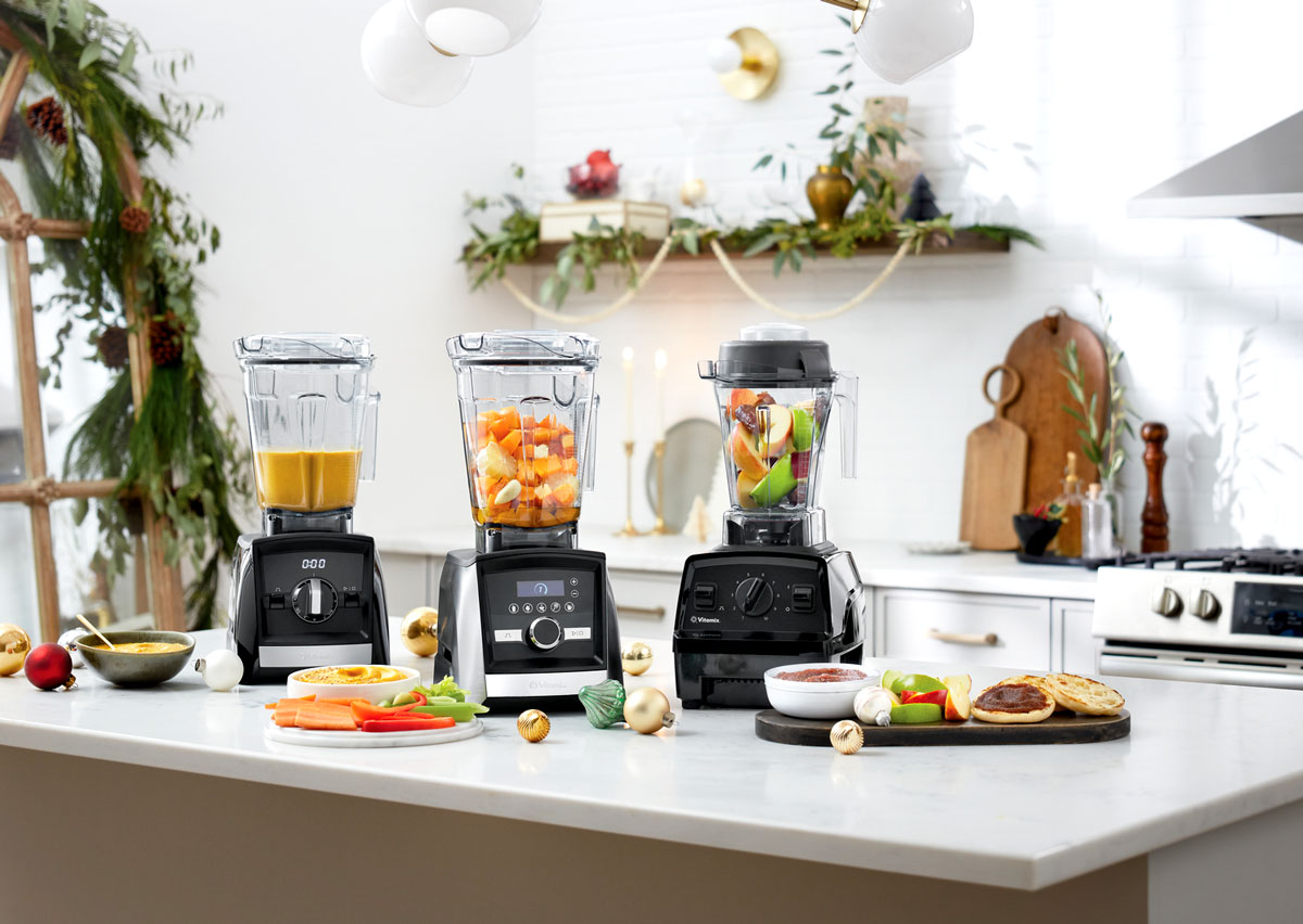 Three vitamix blenders on a light gray countertop with apples and cut up veggies