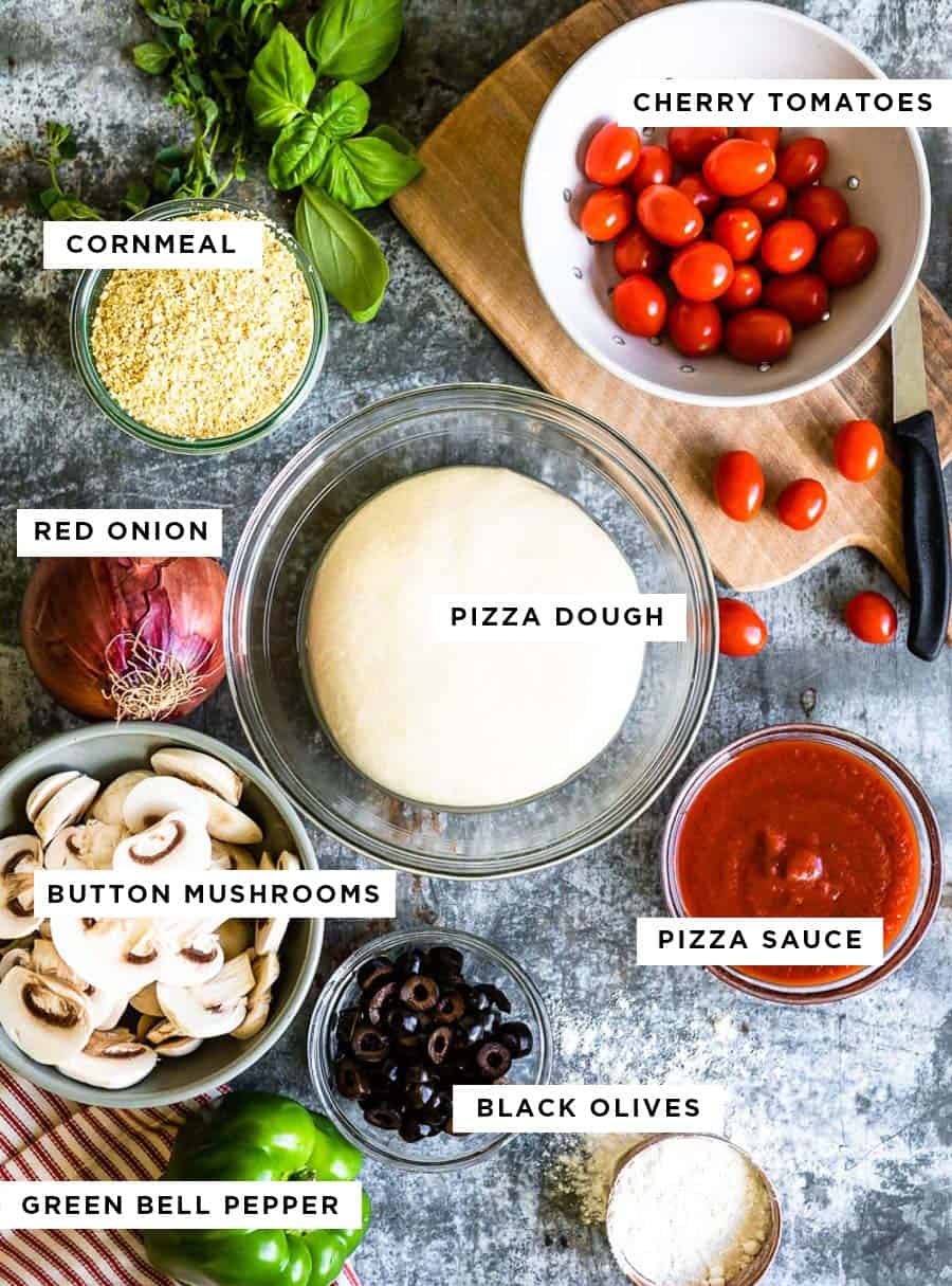 ingredients for homemade pizza including cherry tomatoes, cornmeal, red onion, pizza dough, button mushrooms, pizza sauce, black olives and green bell pepper.