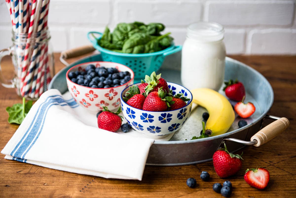 ingredients for a healthy snack including blueberries, strawberries, banana, spinach and plant milk.