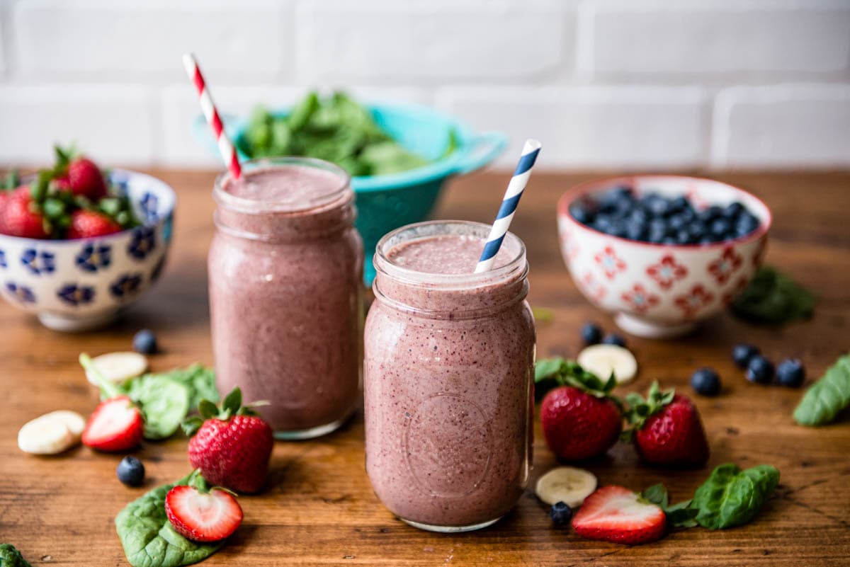 2 mason jars of a berry smoothie with bowls of fresh berries surrounding it.