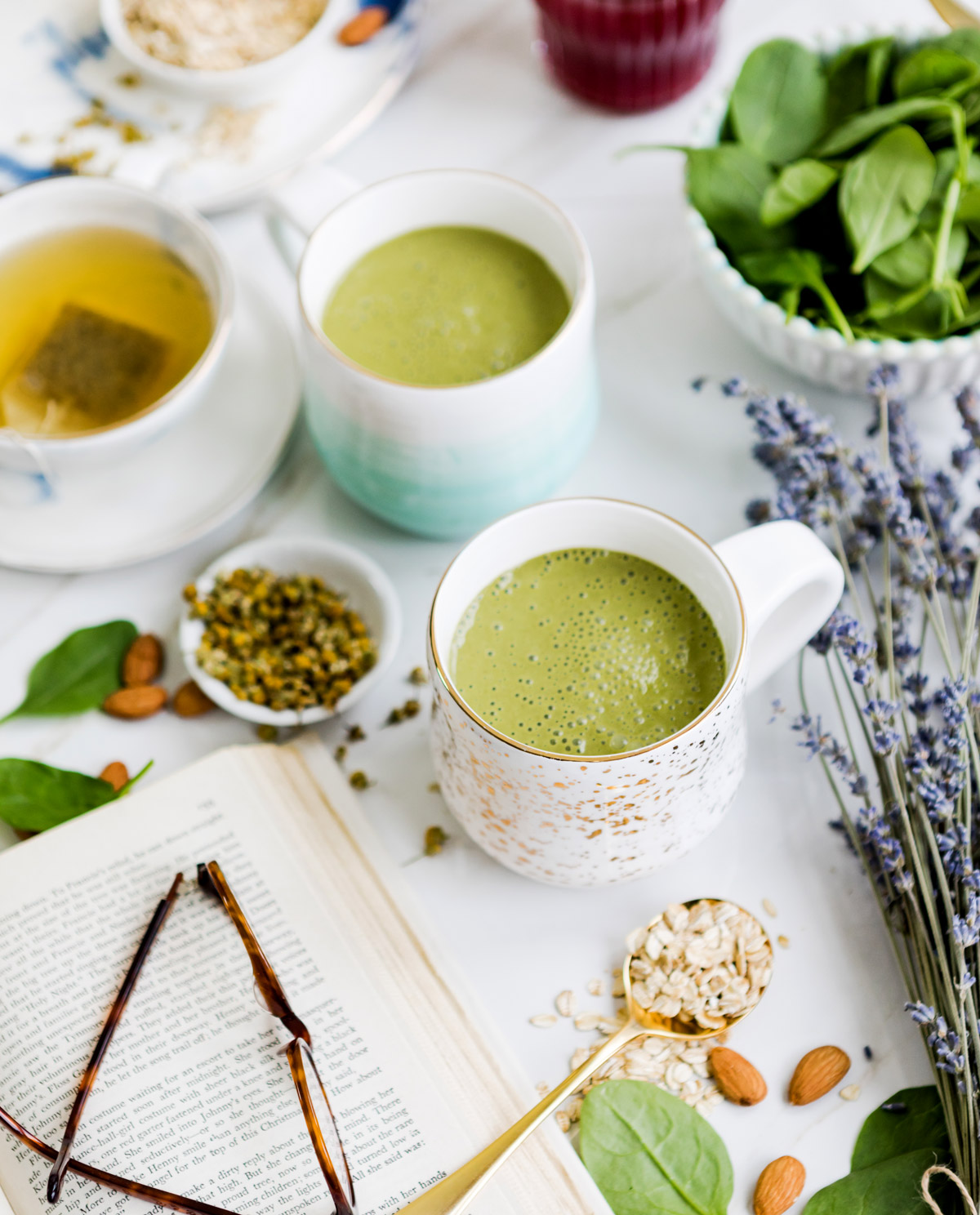 Two mugs of green sleep smoothie surrounded by lavender, spinach, almonds, and a mug of tea highlighting ingredients that promote relaxation before bed.