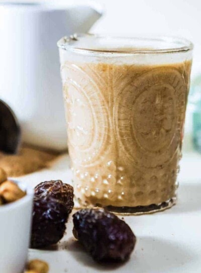 Healthy caramel protein shake in an etched glass next to pitted dates and a white bowl of cashews.