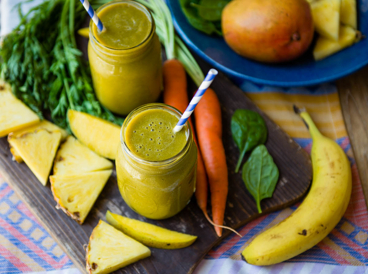 2 glass jars of carrot greens smoothie with papers straws, surrounded by fresh carrots, mango and pineapple slices.