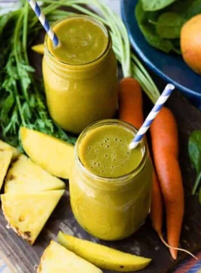 2 glass jars of carrot greens smoothie with papers straws, surrounded by fresh carrots, mango and pineapple slices.