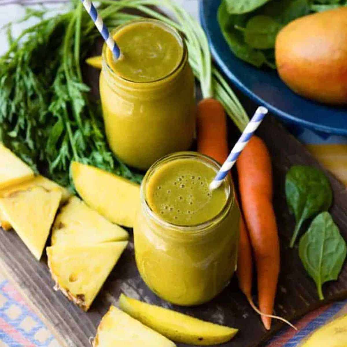 2 glass jars of carrot greens smoothie with papers straws, surrounded by fresh carrots, mango and pineapple slices.