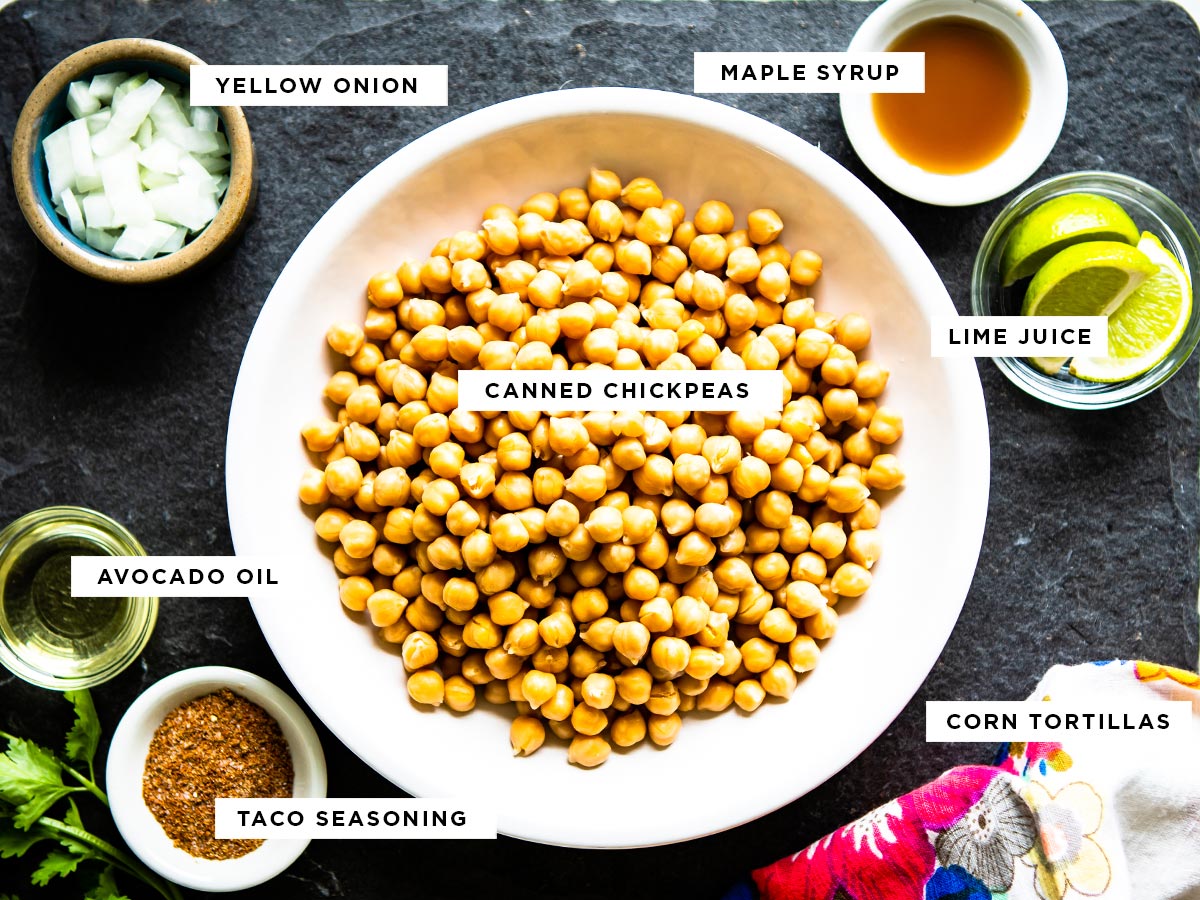 ingredients for vegan chickpea tacos including yellow onion, maple syrup, lime juice, canned chickpeas, corn tortillas, taco seasoning and avocado oil.