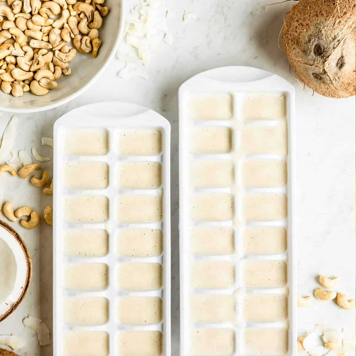 2 white ice cube trays filled with frozen coconut cubes on a white counter top.