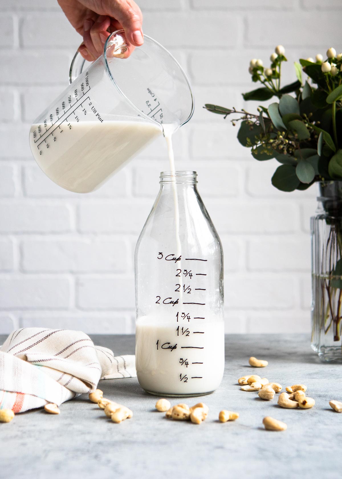 pouring homemade plant based beverage into a pitcher with cashew pieces scattered on the counter.