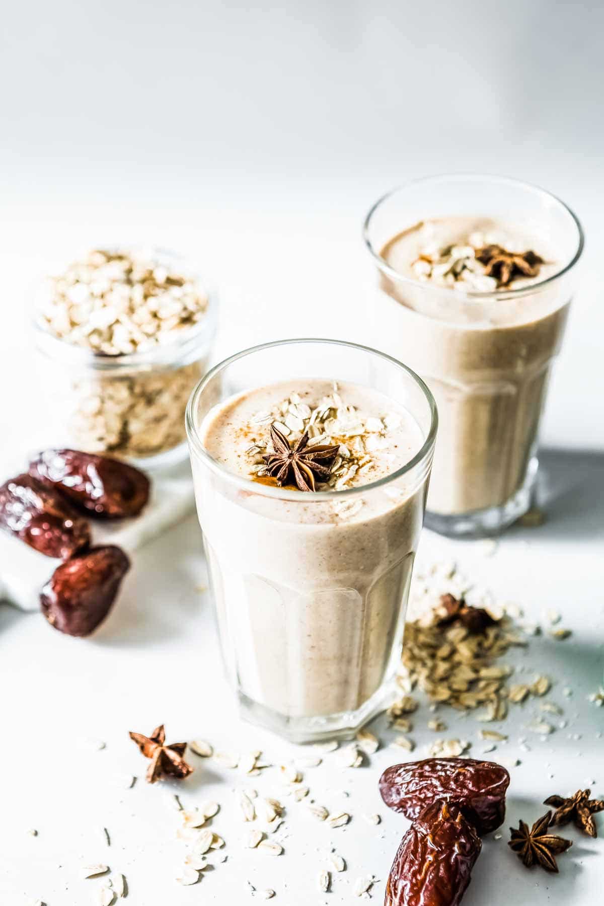 Date smoothie recipe in tall glass cups, garnished with star anise, and served on a white countertop with Medjool dates and oats sprinkled around them. 