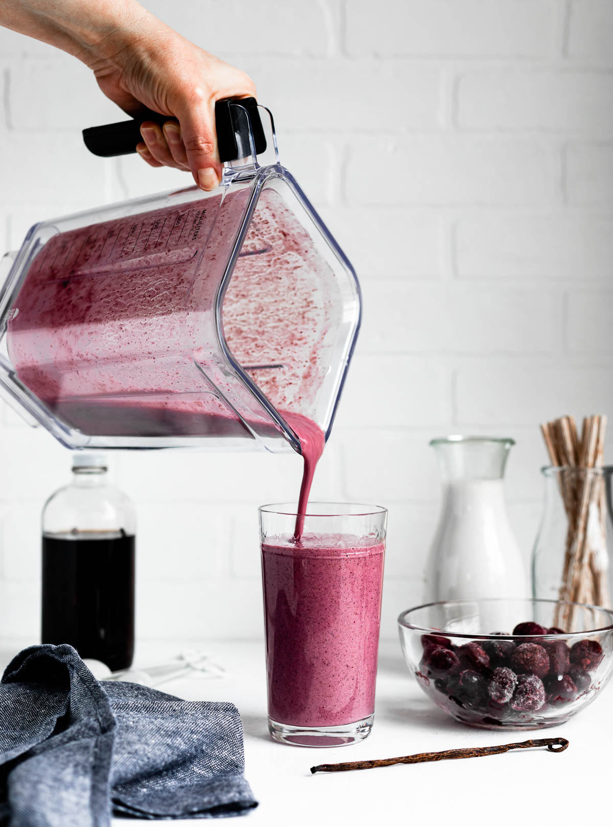 pouring a pink smoothie into a tall glass on a white counter top surrounded by smoothie ingredients.