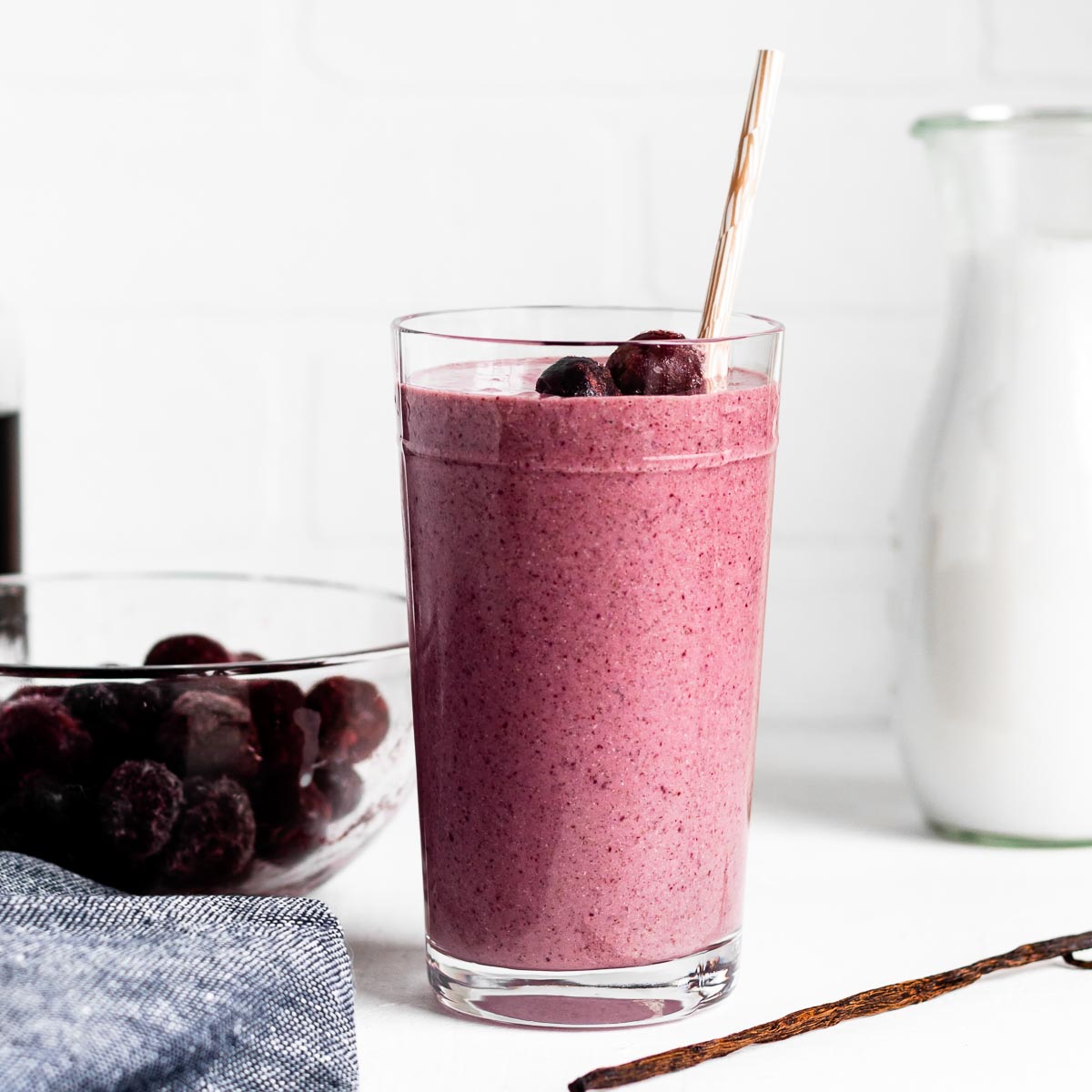 glass with pink diabetes smoothie topped with frozen cherries, a whole vanilla bean and a paper straw.