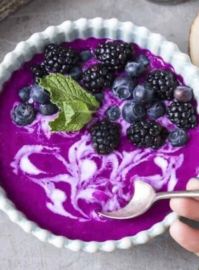 white scalloped bowl with purple dragon fruit smoothie in it topped with blueberries, blackberries, coconut whipped cream and a spoon.