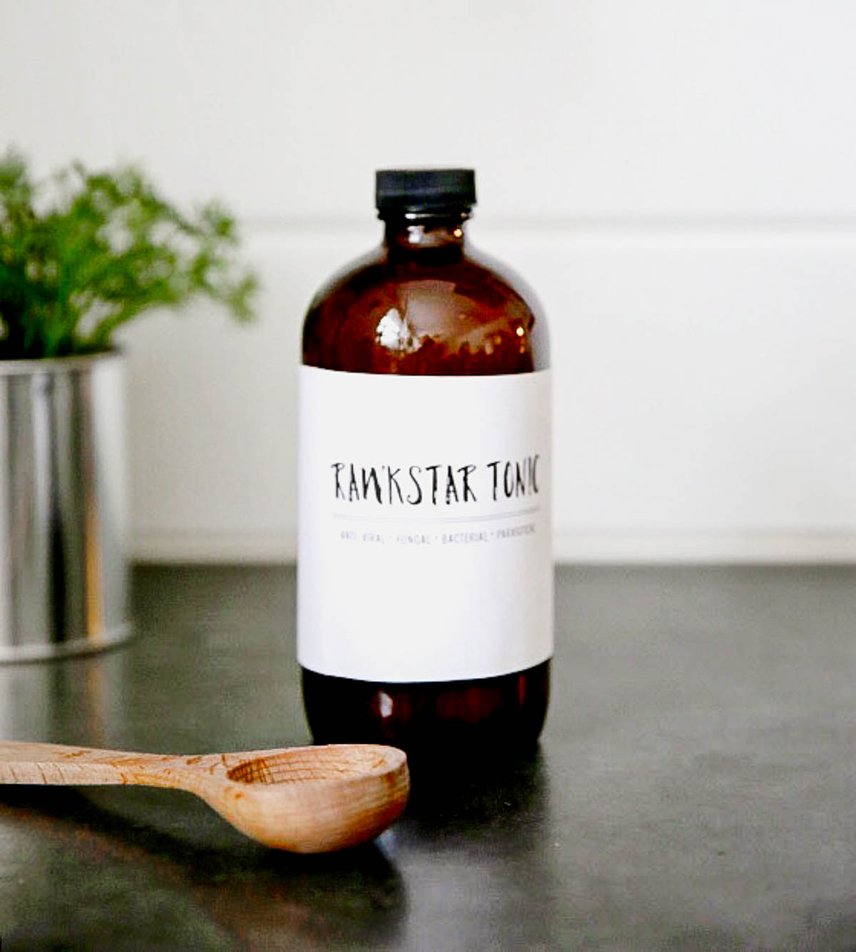 dark brown glass jar with white label reading Rawkstar Tonic, a fire cider recipe on a table with a wooden spoon.