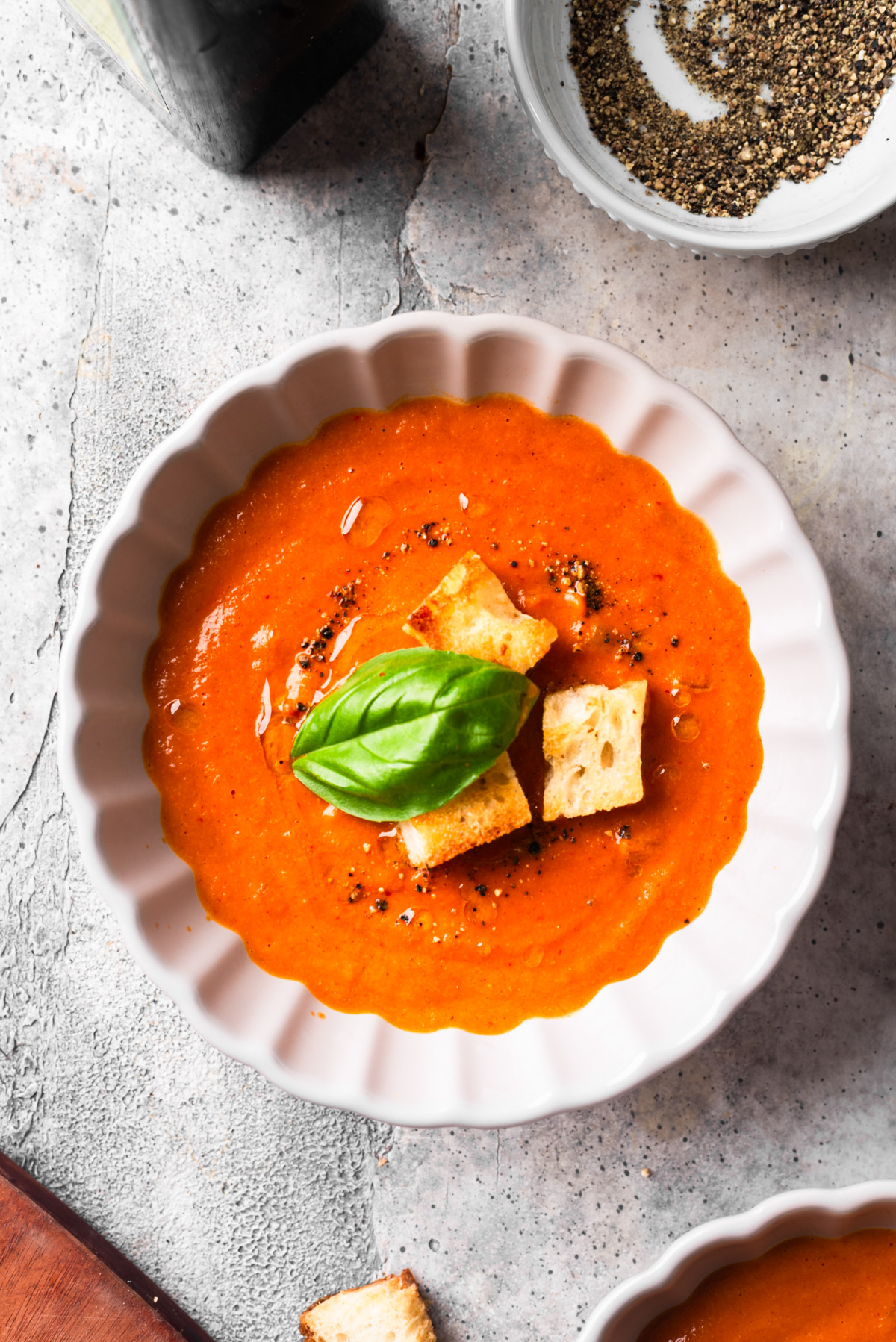 Big bowl of fire-roasted tomato soup with fresh basil and croutons on gray table.