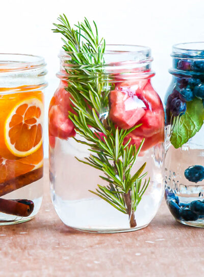 3 glass jars of fruit-infused water with ingredients like oranges, cinnamon sticks, rosemary, strawberries, blueberries and basil.