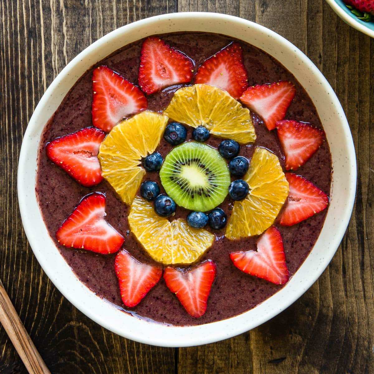 white bowl of smoothie topped with fresh fruit like strawberries, oranges, blueberries and kiwi.