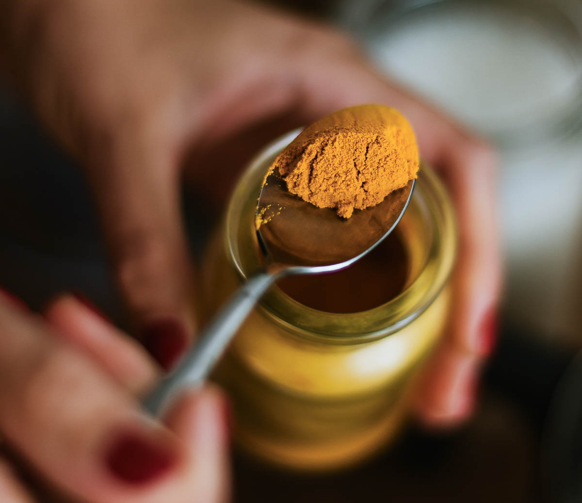 2 hands scooping out ground turmeric from a glass jar using a silver spoon.