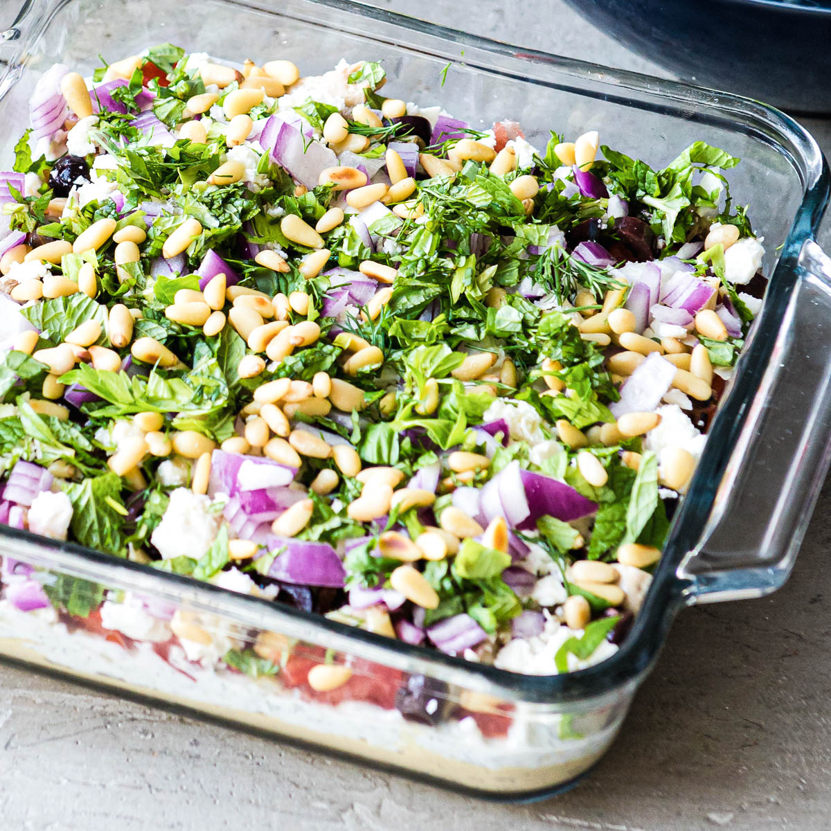 glass dish filled with layered Greek dip including fresh parsley, purple onions, white beans and olives.