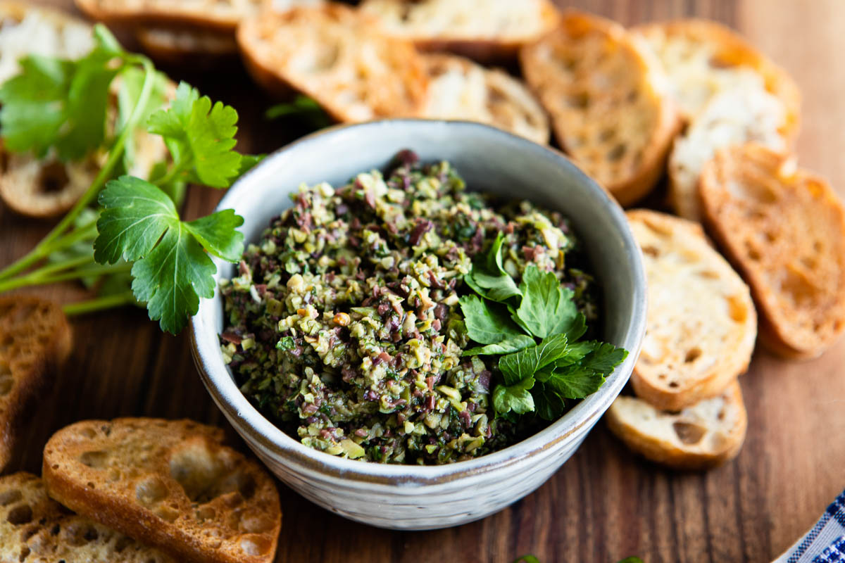 green olive tapenade in a bowl topped with fresh parsley, surrounded by crostini.