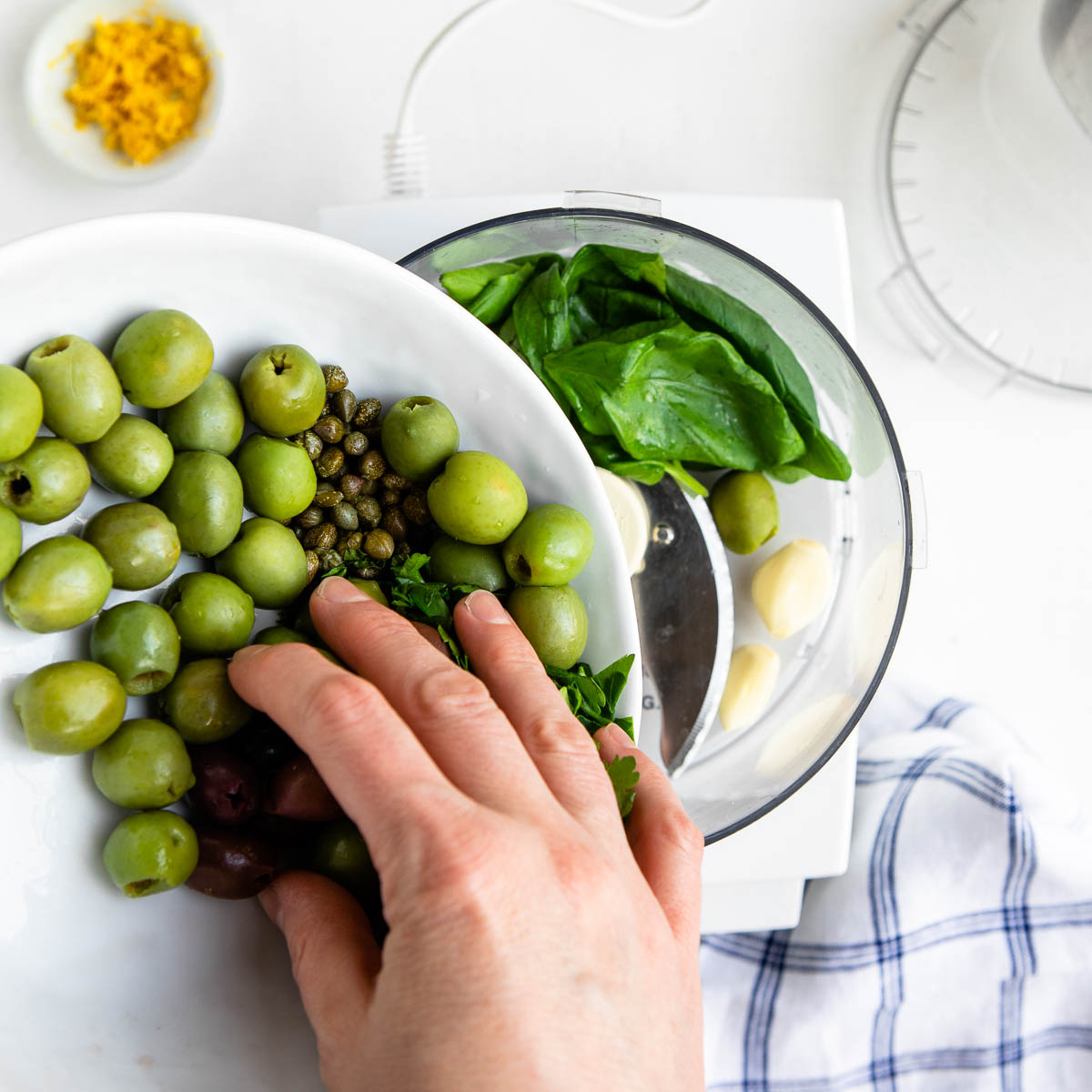 a hand grabbing a plate full of green olives, cappers and herbs for a green olive tapenade. 