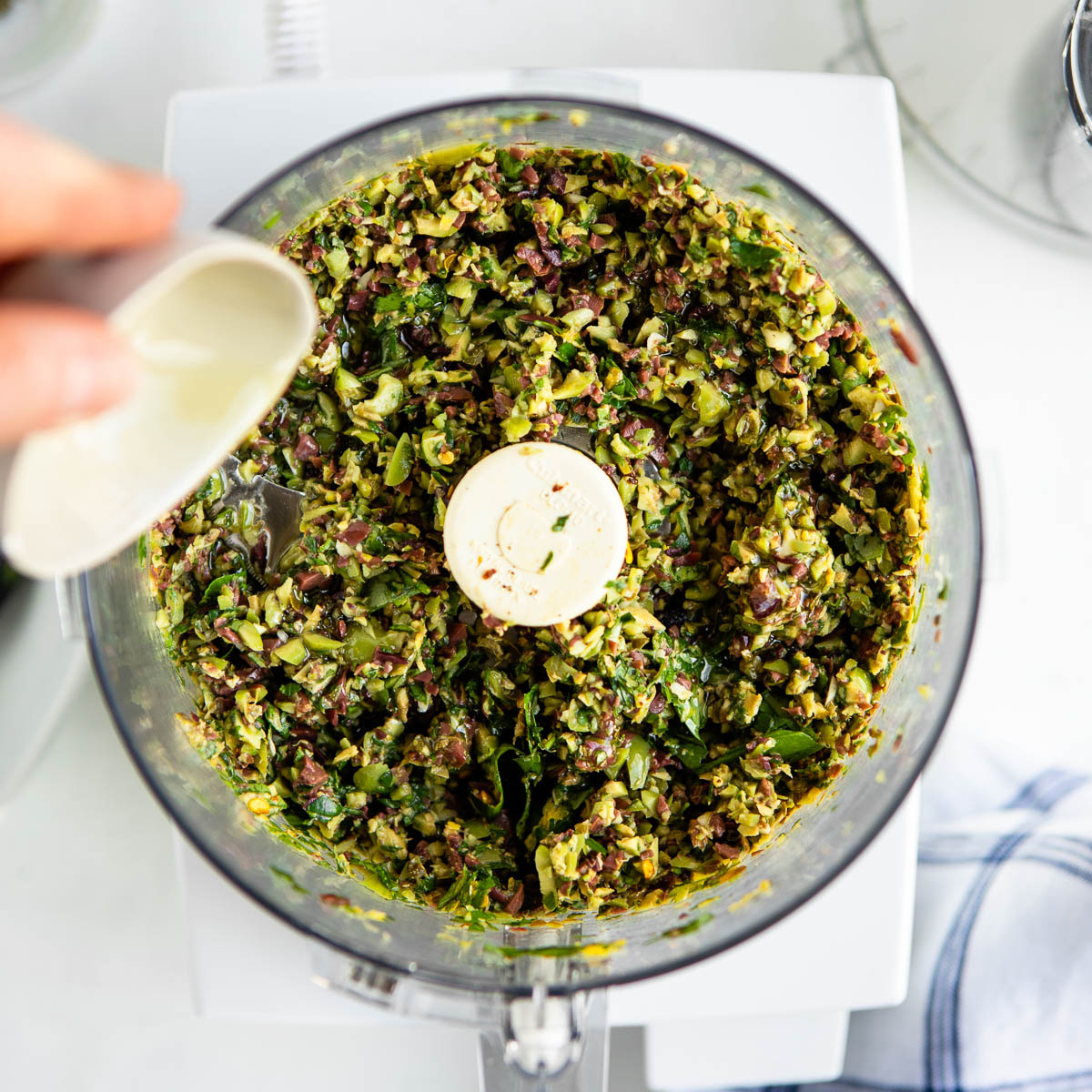 chopped up ingredients in a food processor and a hand pouring in olive oil from a small white bowl. 