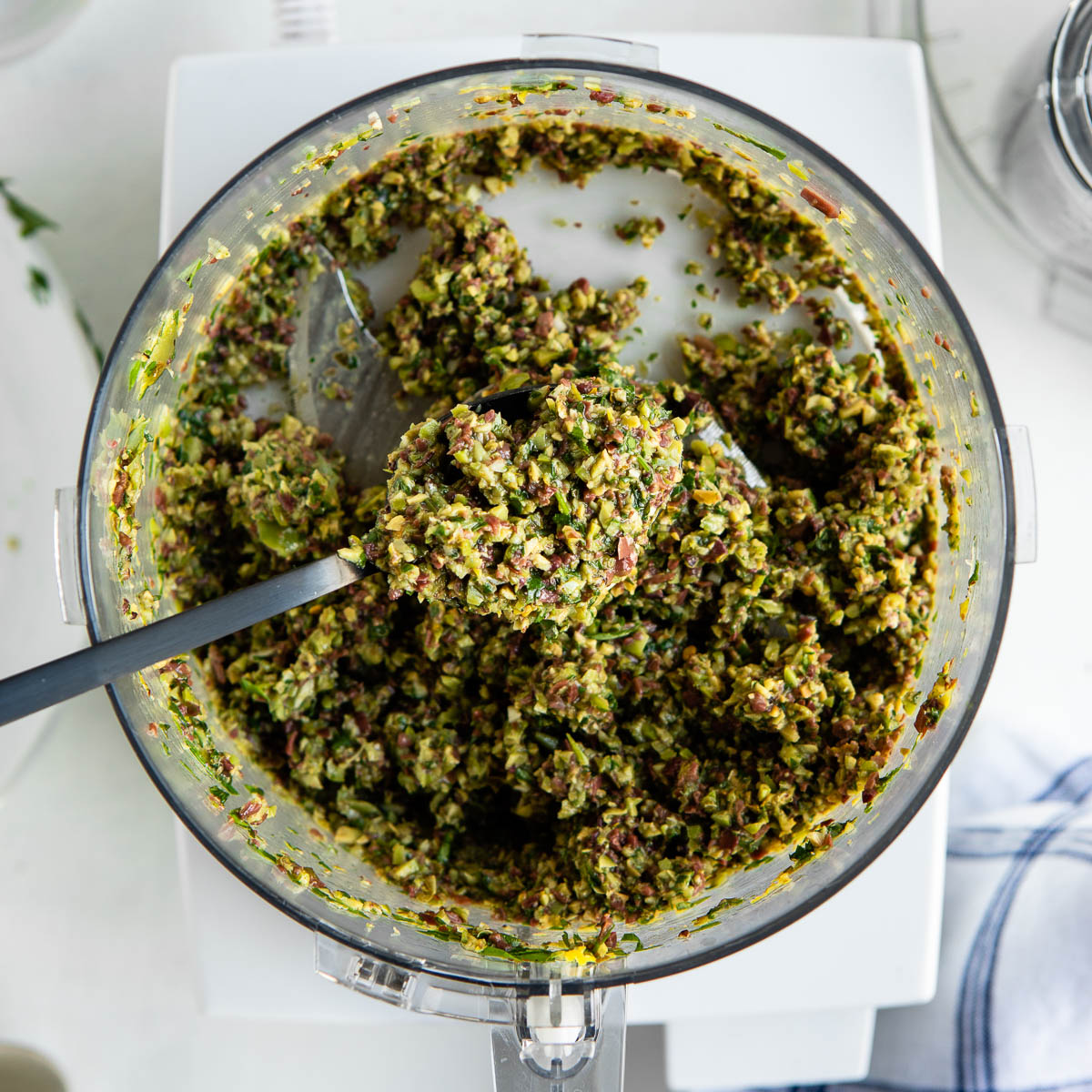 a black spoon scooping olive tapenade out of the bowl of a food processor.