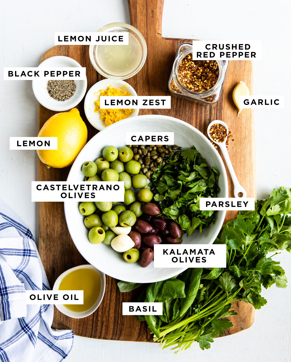 labeled ingredients for green olive tapenade including lemon juice, crushed red pepper, black pepper, lemon zest, garlic, lemon capers, castelvetrano olives, kalamata olives, parsley, olive oil and basil.