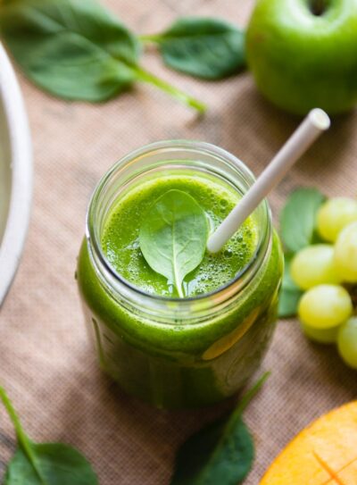 Green tea smoothie in a glass mason jar with a paper straw, surrounded by grapes, mango, apple and spinach.