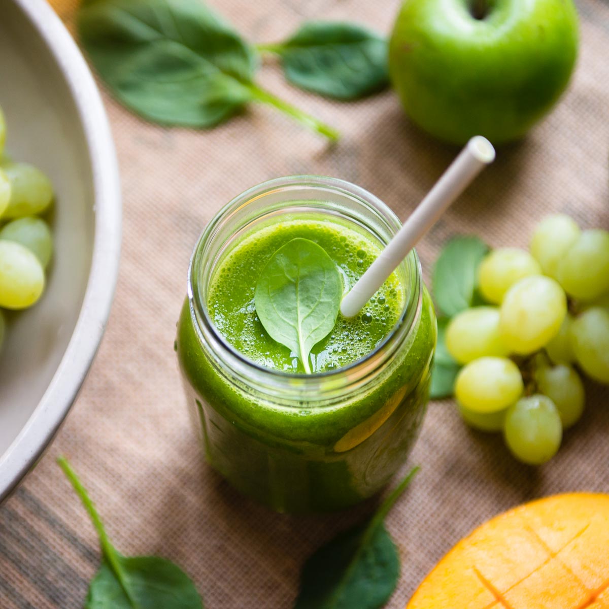 Green tea smoothie in a glass mason jar with a paper straw, surrounded by grapes, mango, apple and spinach.