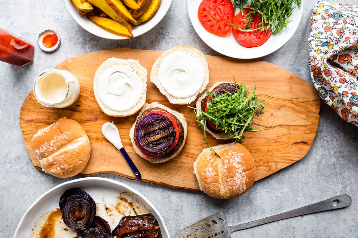 grilled vegan burgers with fresh toppings on a wooden board next to a glass jar of garlic aioli.