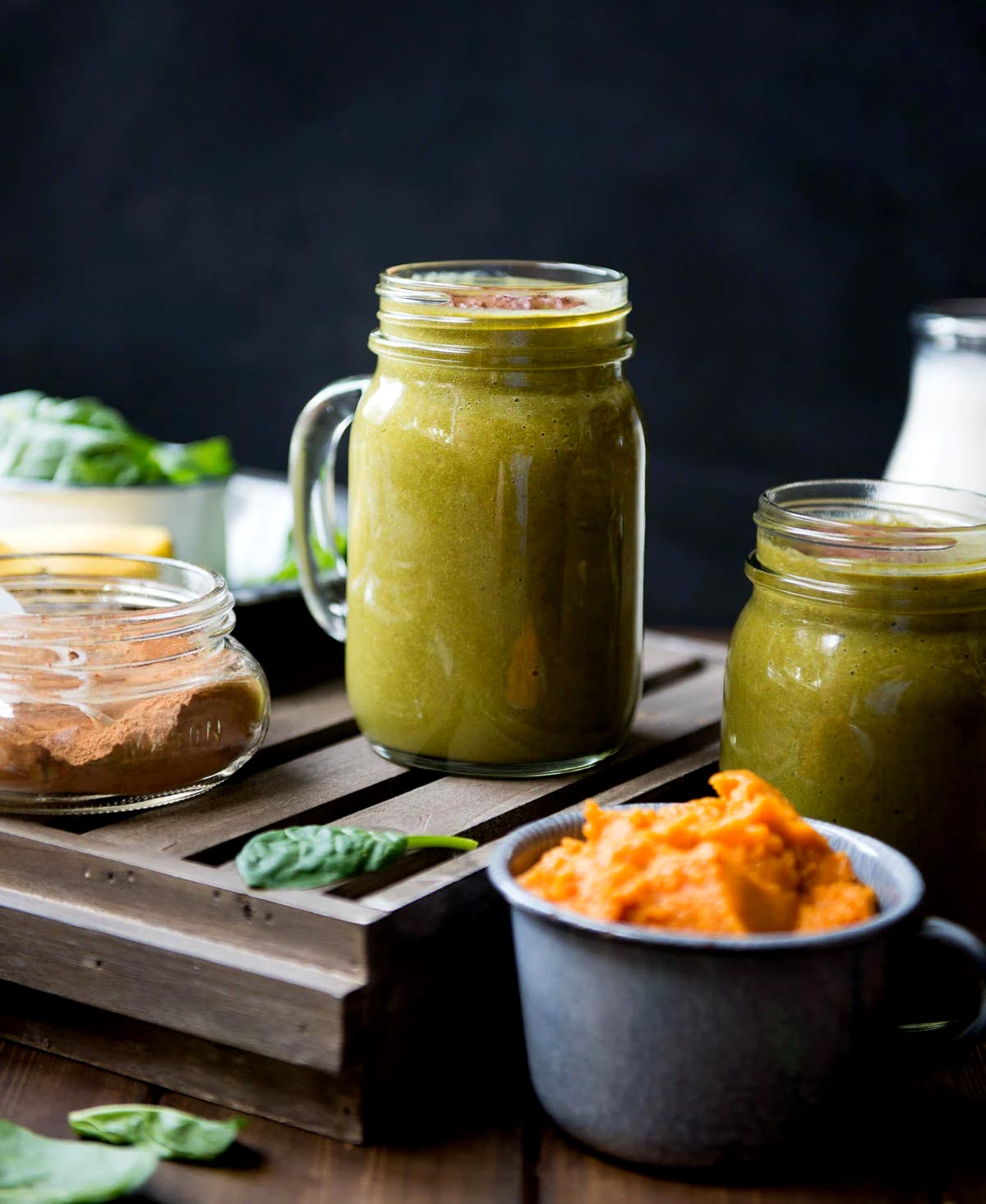 Two glass mug of pumpkin pie smoothies on a wooden tray next to a jar of spice and a tin cup of pumpkin puree.