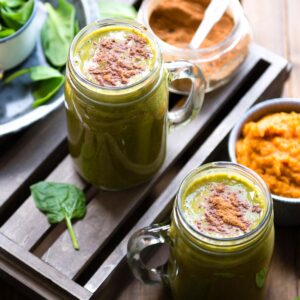 Glass mugs with pumpkin smoothie topped with pumpkin pie spice next to a mug of pumpkin puree and a jar of spice.