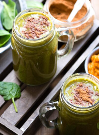 Glass mugs with pumpkin smoothie topped with pumpkin pie spice next to a mug of pumpkin puree and a jar of spice.