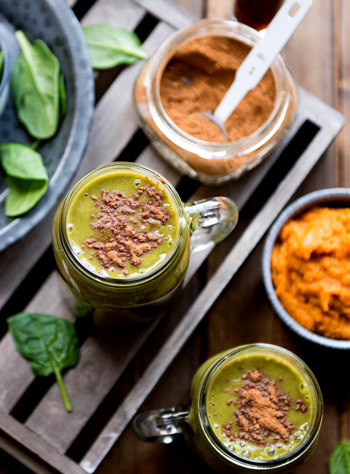 Two glass jars of pumpkin smoothie topped with pumpkin pie spice, next to a mug of pumpkin puree, a jar of spice, and blurred ingredients. 