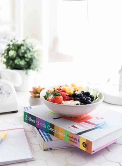 white bowl of fruits and nuts on top of 2 books on a white desk.